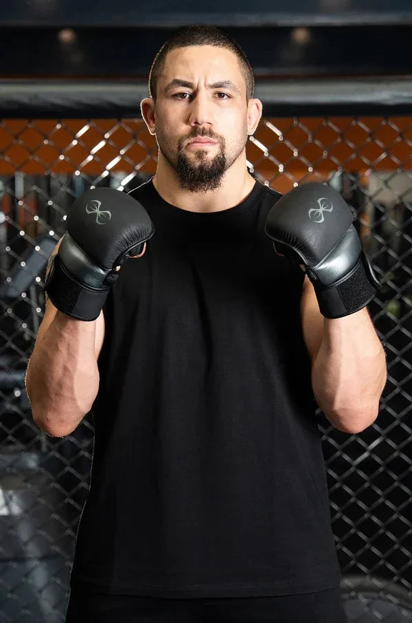 Man in black boxing gloves and sleeveless shirt poses in front of gym cage