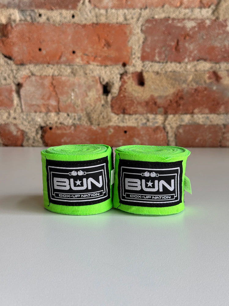 Pair of neon green boxing hand wraps with black and white BUN logo on a table, brick wall background