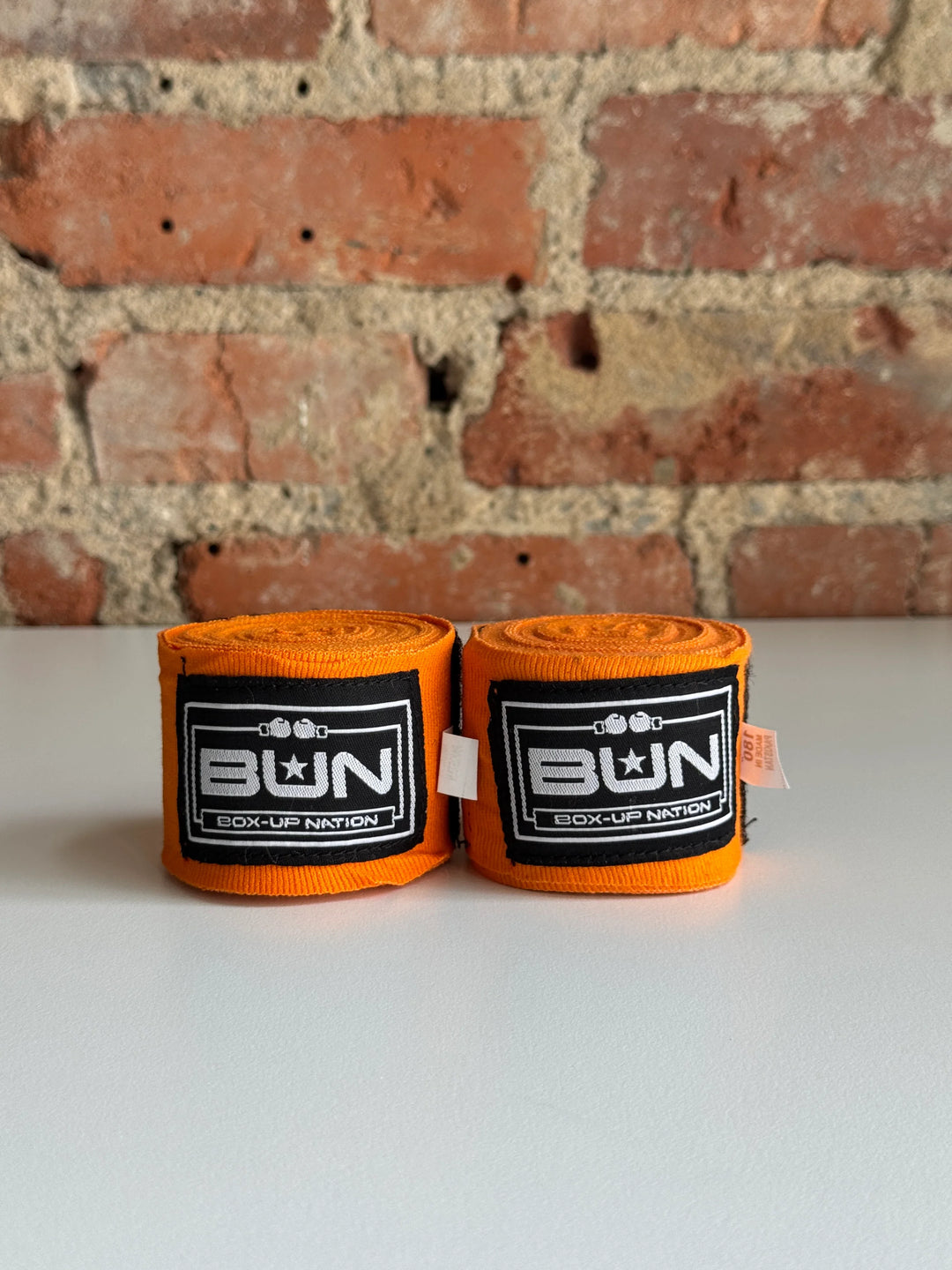 Pair of orange boxing hand wraps with black Box-Up Nation logo on white table, brick wall background.