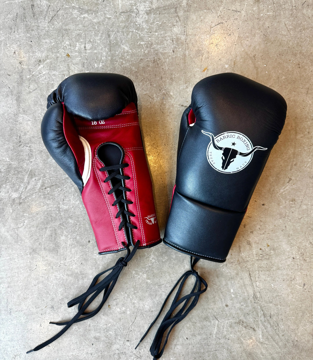 Red and black lace-up boxing gloves with Barric Boxing Texas logo on concrete floor