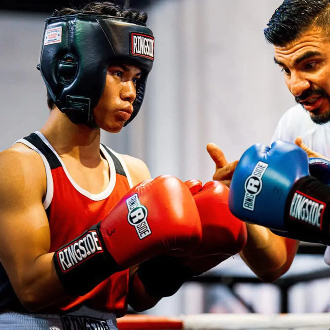 Boxer in headgear and red gloves receiving coaching in boxing gym, Ringside equipment visible.